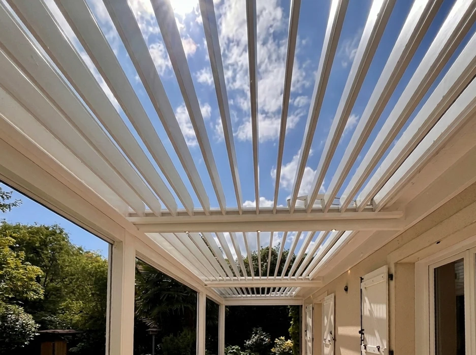 Terrasse couverte vue de l'intérieur, pergola bioclimatique à lames orientables en aluminium blanc, position ouverte laissant apparaître un ciel bleu nuageux