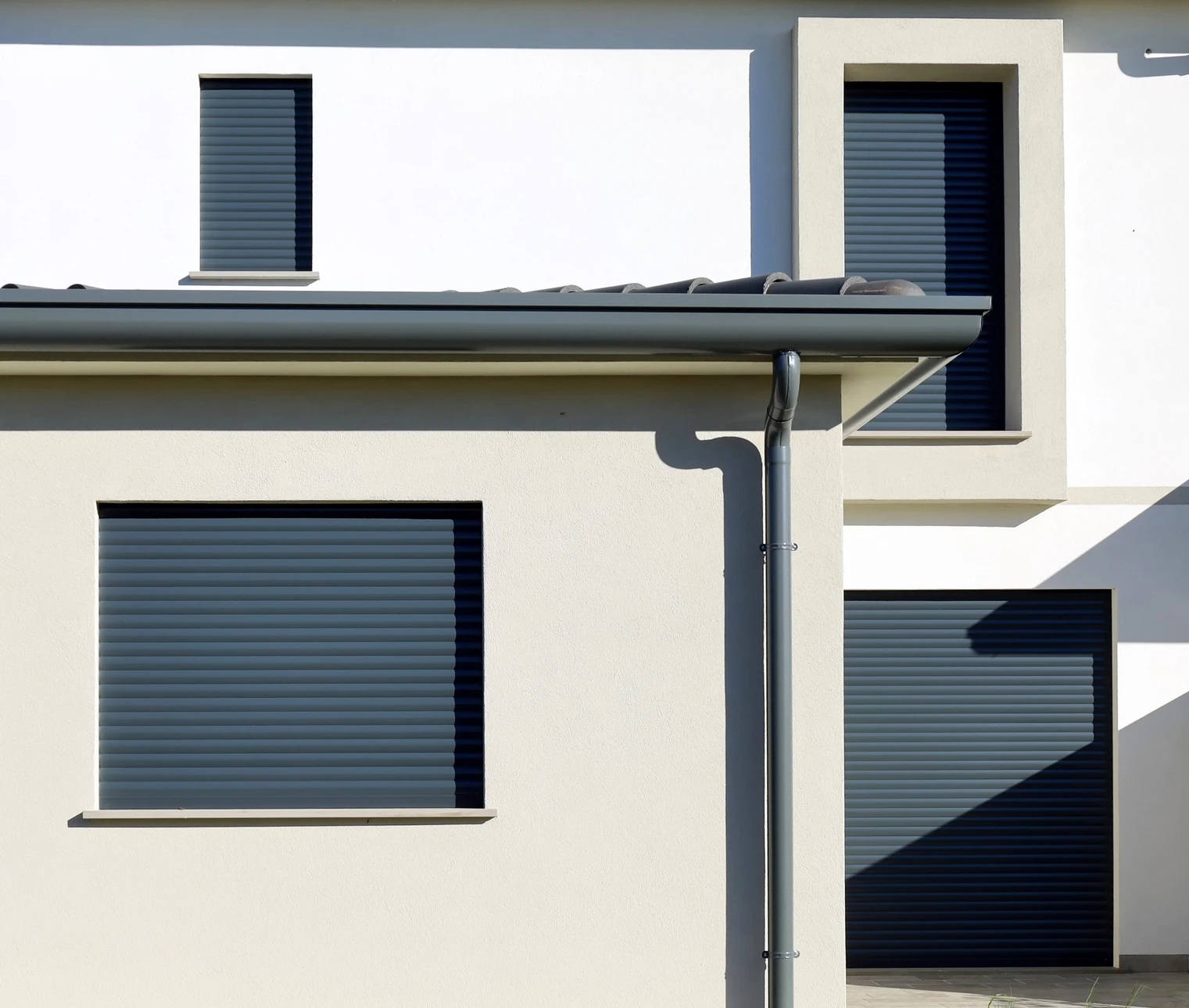 Façade moderne d'habitation avec volets roulants gris fermés, murs crépis beige clair, gouttière et descente pluviale anthracite