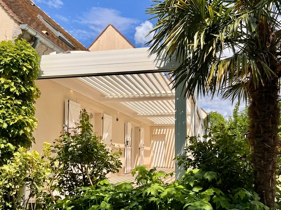 Pergola bioclimatique à lames orientables blanches, fixée en mur sur une maison de style français