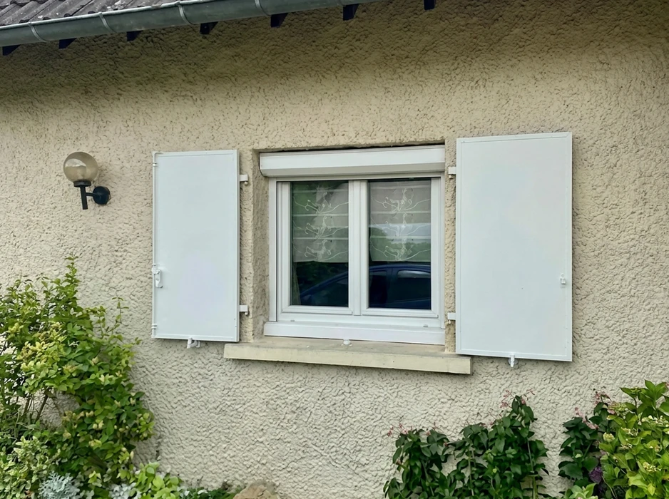 Façade de maison en enduit texturé beige avec une fenêtre PVC blanche à deux vantaux coulissants, équipée d'un volet roulant coffre apparent