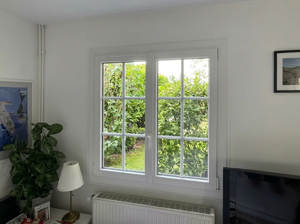 Pièce intérieure lumineuse et confortable d'une maison rénovée, vue depuis l'intérieur avec une grande baie vitrée ou fenêtre PVC blanc moderne offrant une vue claire sur le jardin ensoleillé. La lumière naturelle inonde la pièce, révélant un intérieur chaleureux et bien aménagé. Pas de personnes. L'accent est mis sur la clarté, la luminosité et le sentiment de bien-être procuré par une menuiserie de qualité.