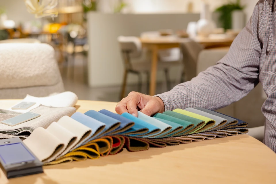 Intérieur professionnel avec personne présentant éventail d'échantillons textiles colorés dans une main, piles de tissus d'ameublement neutres disposées sur table