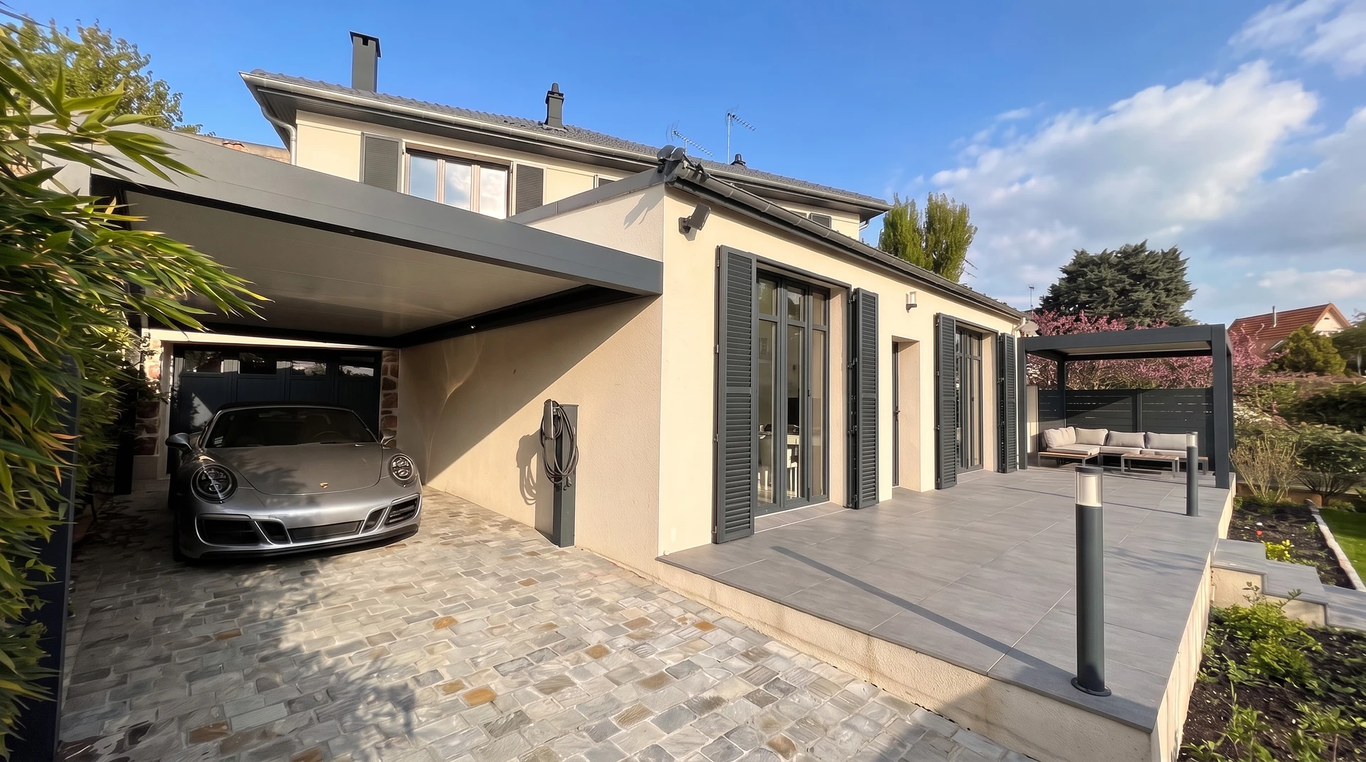 Structure de carport moderne en aluminium blanc, adossée à une maison contemporaine, abritant une voiture, avec lames orientables ajourées permettant une circulation d'air optimal et une luminosité contrôlée. Vue d'ensemble de la structure couverte depuis la cour de la maison, montrant l'abri automobile fonctionnel et épuré en matériau léger.